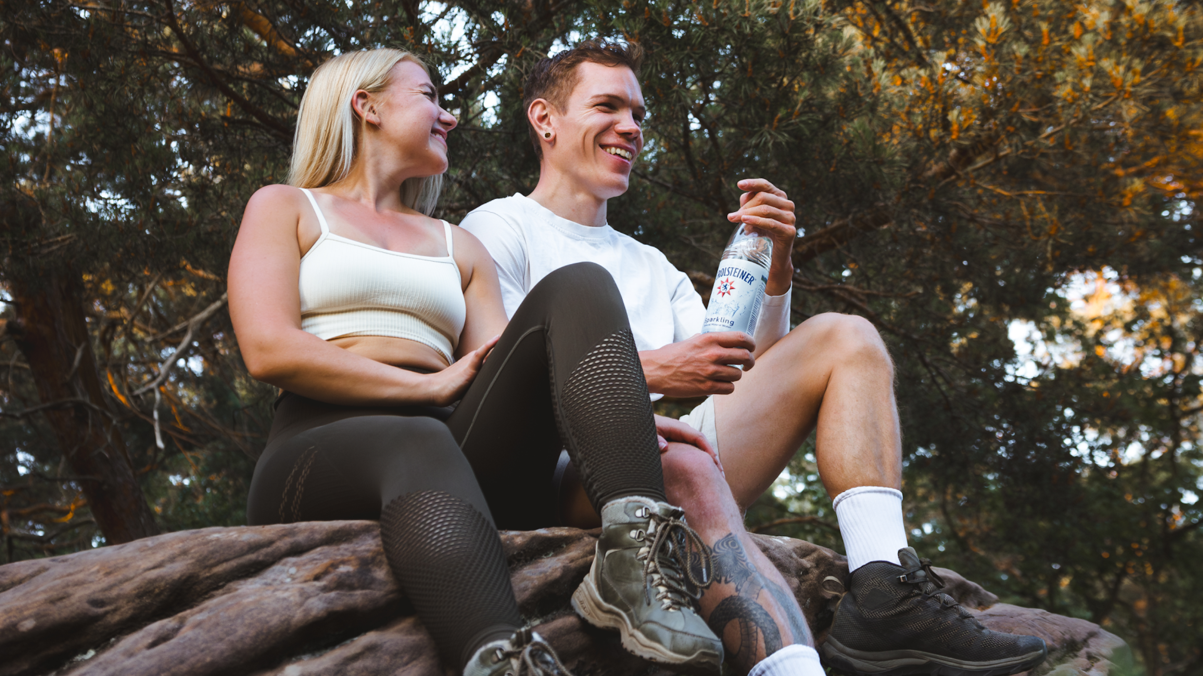 Two people are sitting on a tree trunk in the forest, smiling and holding a bottle of sparkling water.