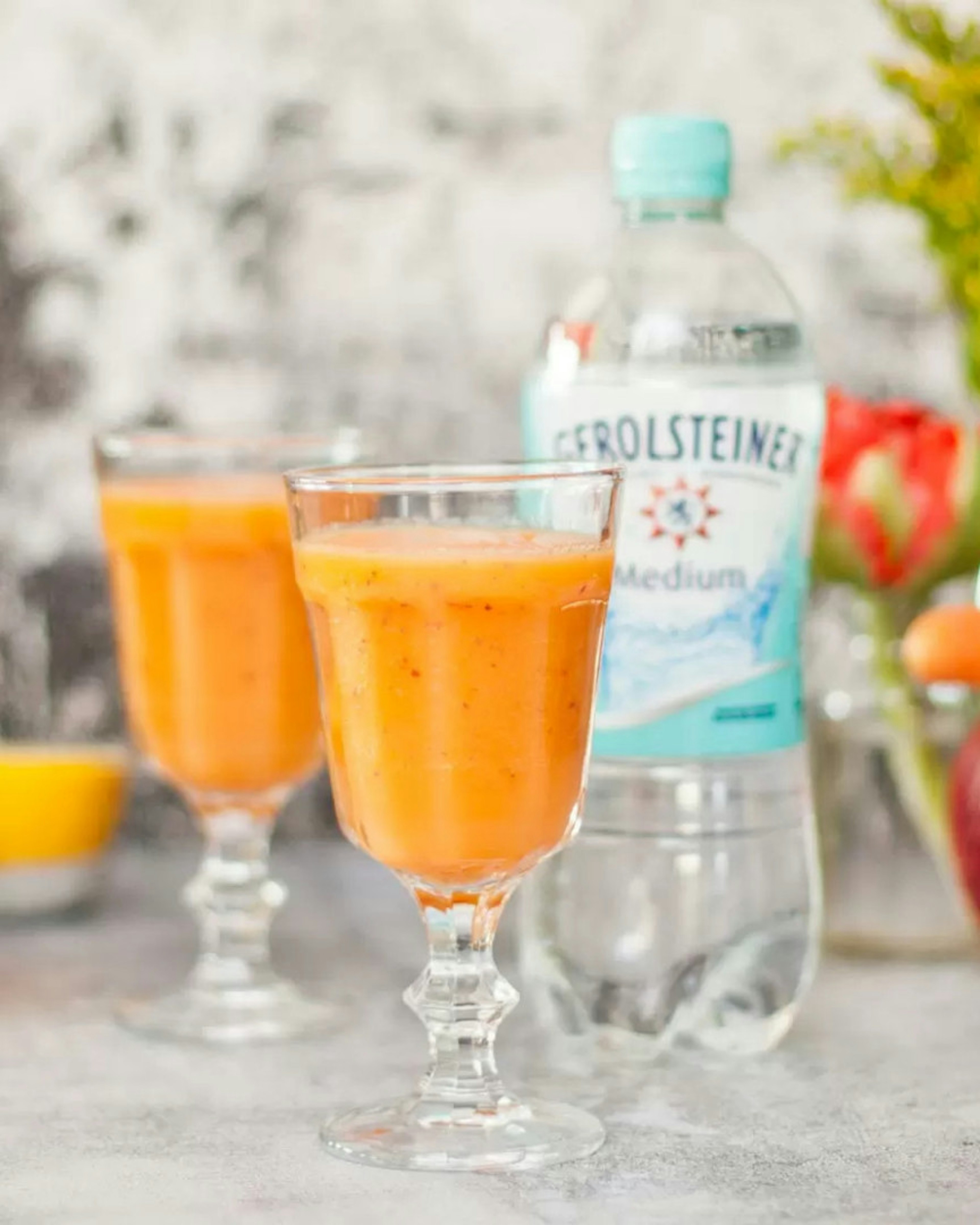 "2 Gläser mit orangenem Smoothie und  Gerolsteiner und Apfel im Hintergrund"