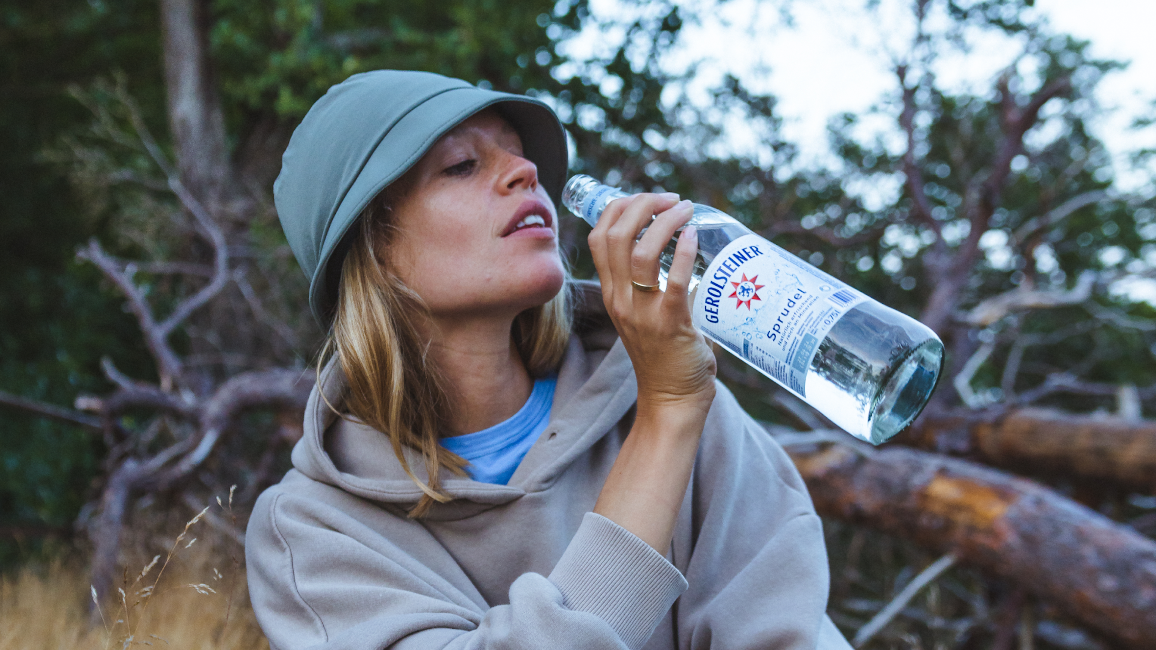 Eine Frau in Outdoor-Kleidung trinkt aus einer Flasche Sprudelwasser in einem Waldgebiet.