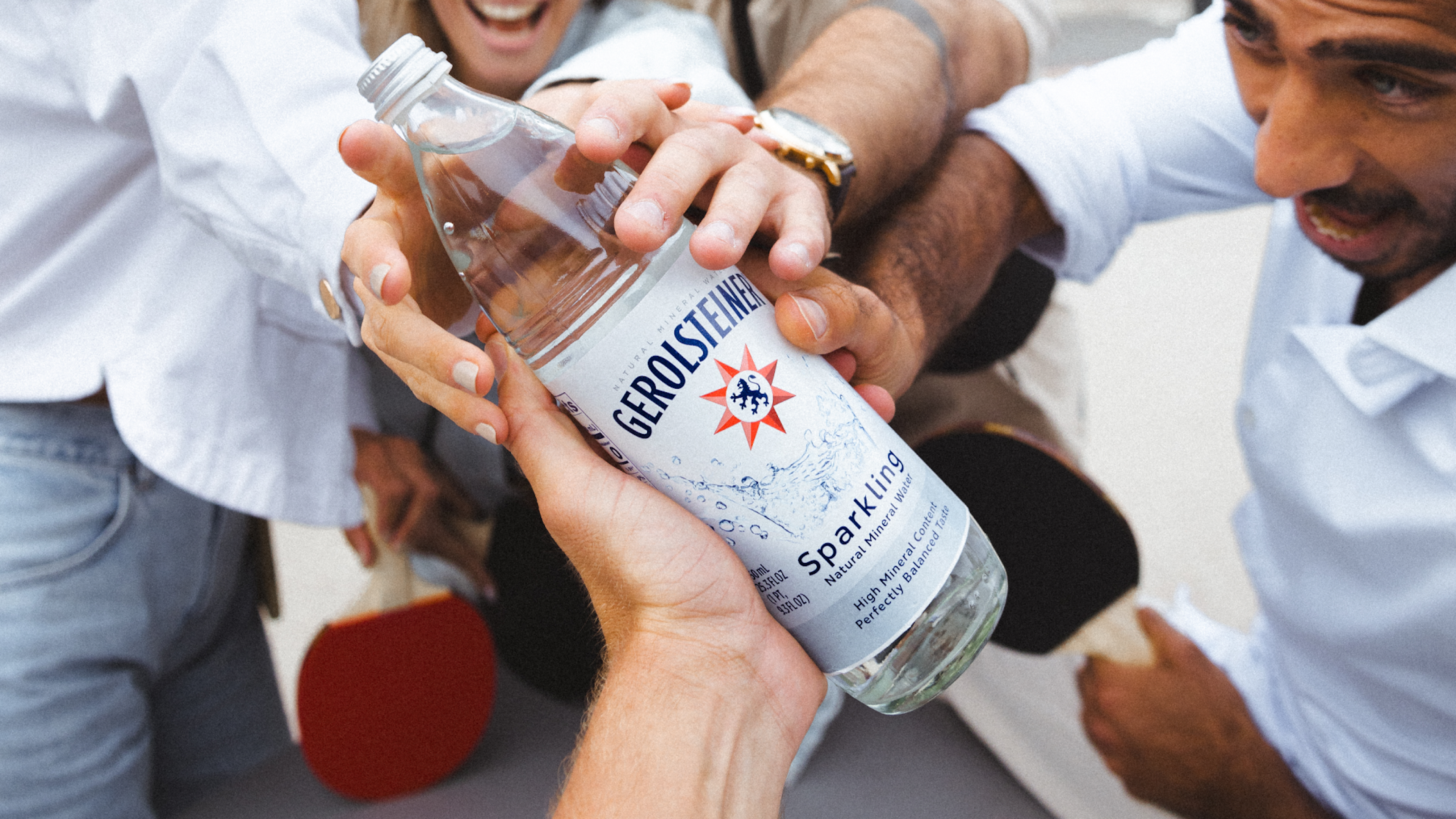 A group of people reach for a bottle of Gerolsteiner sparkling water.