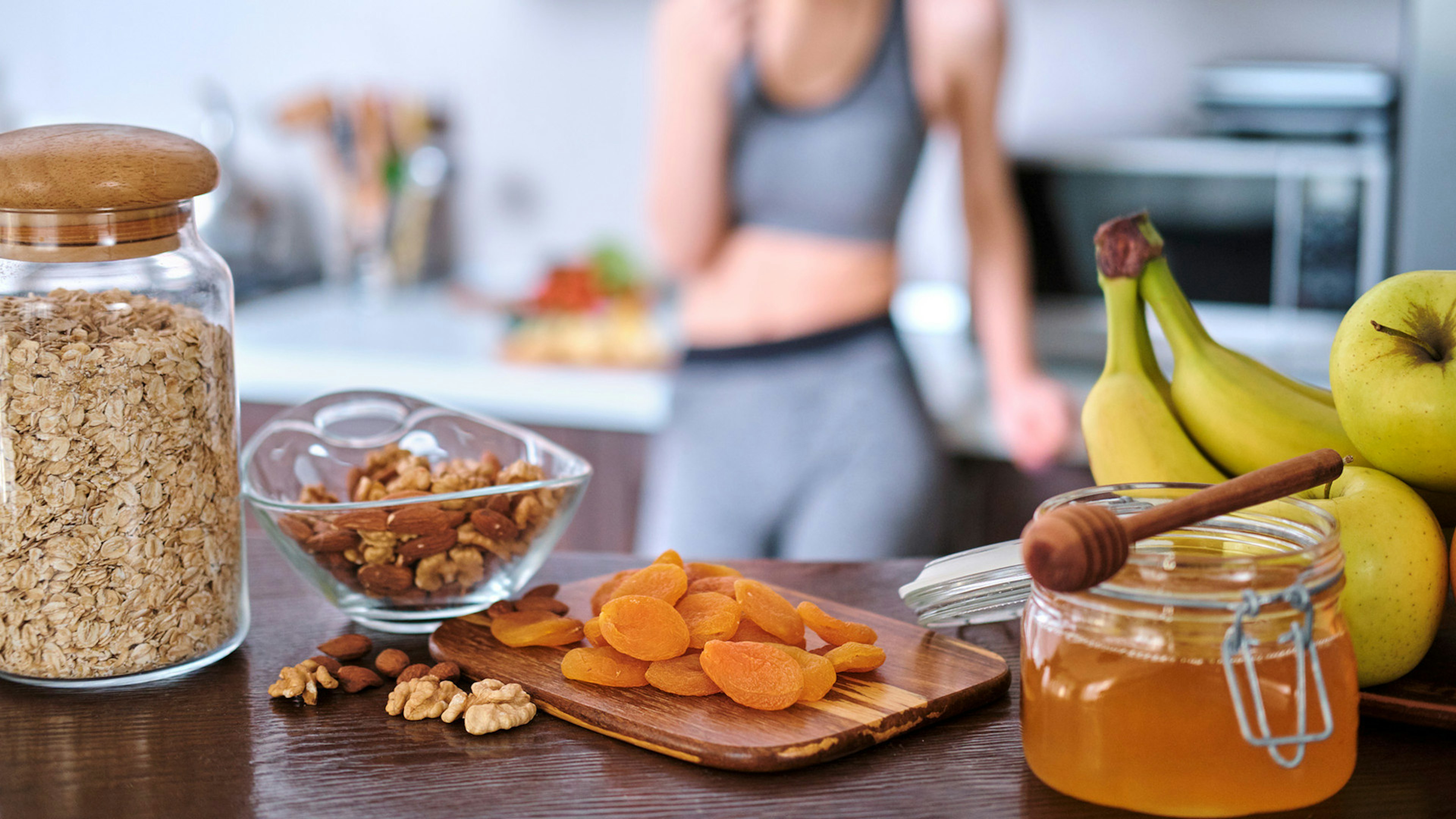 Im Vordergrund stehen gesunde Lebensmittel auf einem Tisch, im Hintergrund isst eine Frau in Sportkleidung einen Apfel.