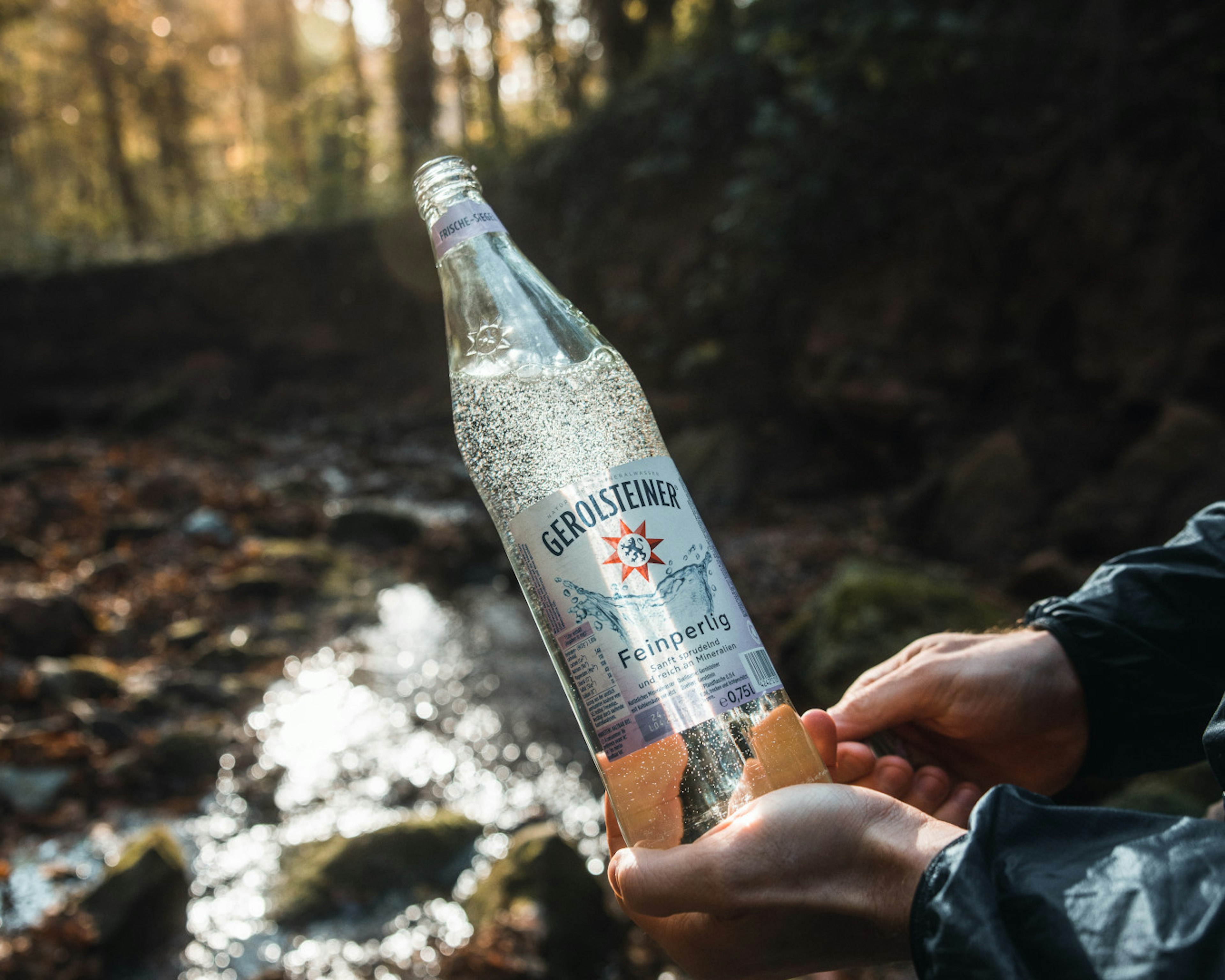 Eine Person hält eine Gerolsteiner Glasflasche der Sorte Feinperlig ins Bild. Die Location ist im Wald an einem Bach