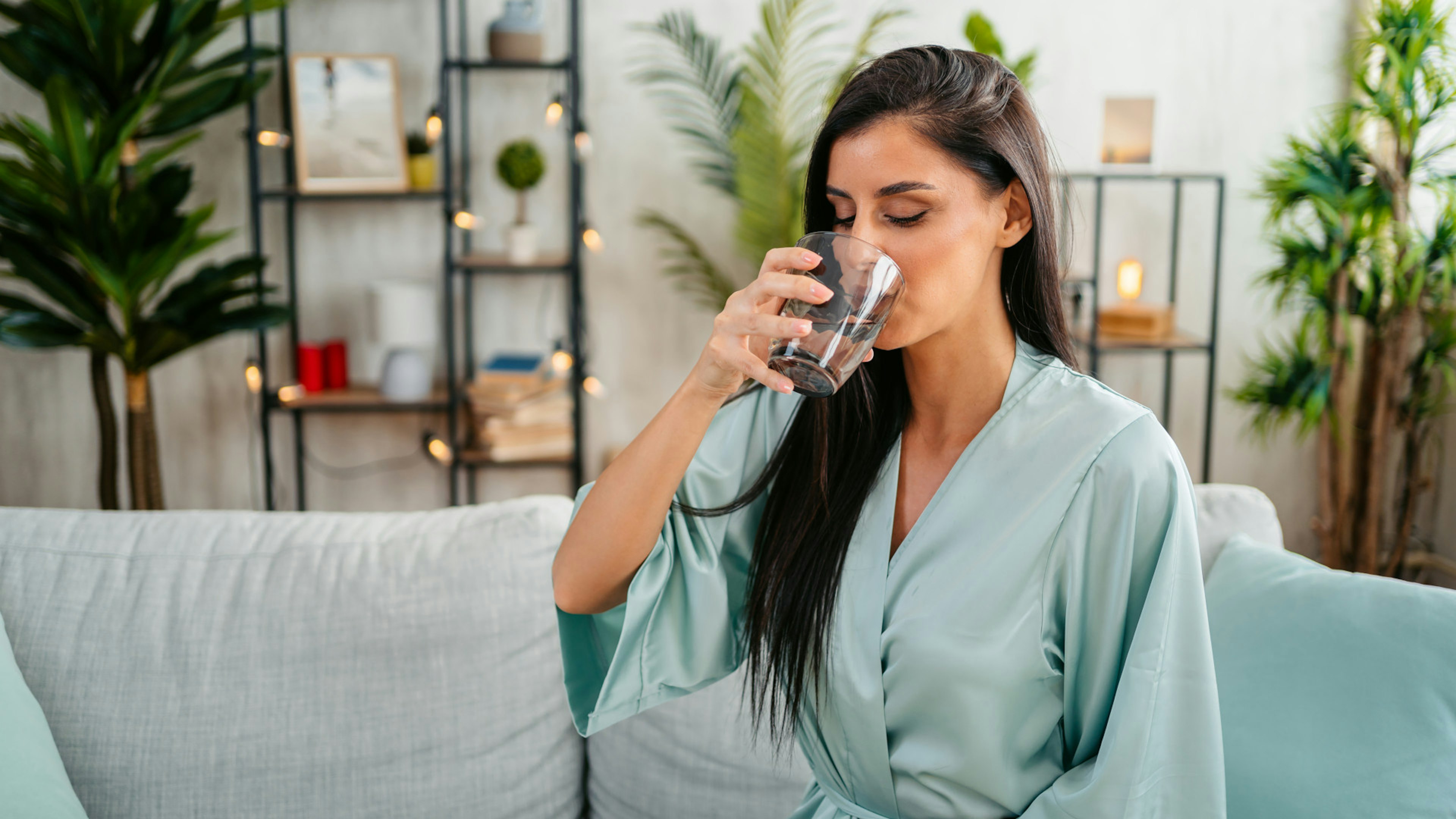 Eine Frau im Bademantel trinkt ein Glas Wasser auf ihrer Couch