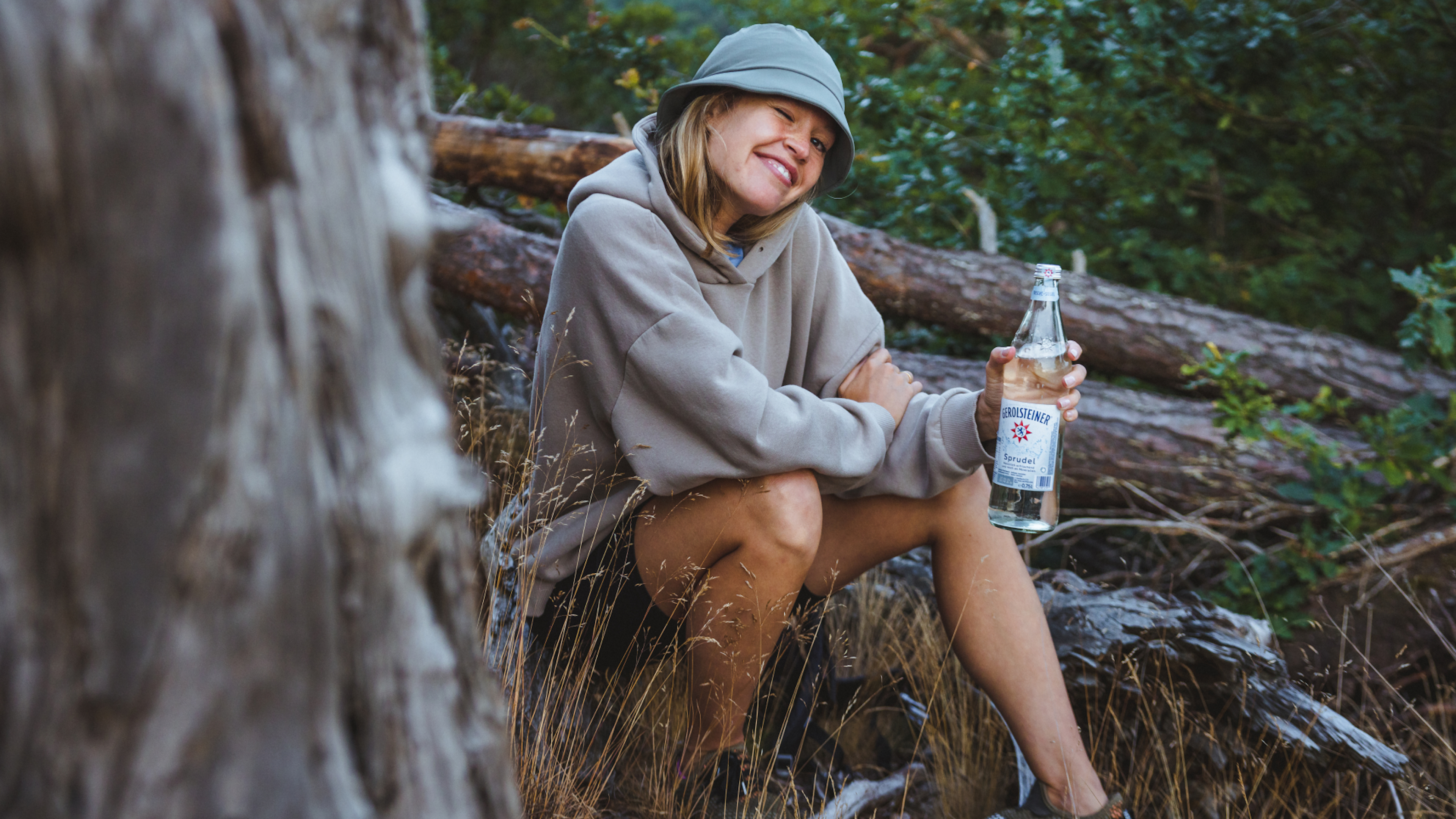 A woman sits on a tree trunk and smiles with a bottle of Gerolsteiner in her hand. 